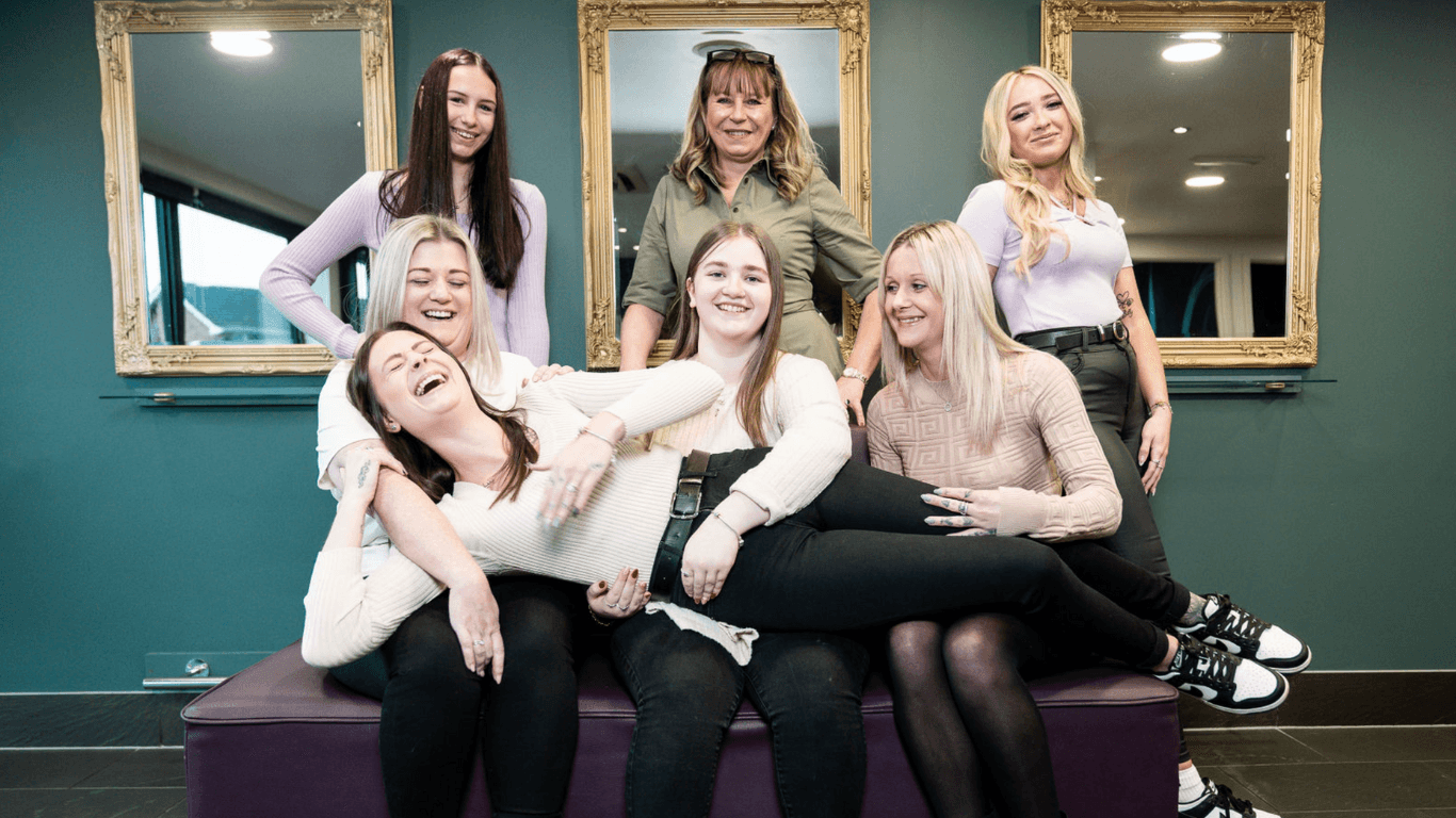 A joyful team photo of the Honeys Hair & Beauty salon in Taunton. Seven women—stylists and therapists—are gathered in front of three gold-framed mirrors on a teal feature wall. The team exudes warmth and camaraderie, with some sitting on a salon bench and one playfully lying across the others’ laps, all smiling and laughing. The image reflects the salon’s friendly, professional, and welcoming atmosphere.