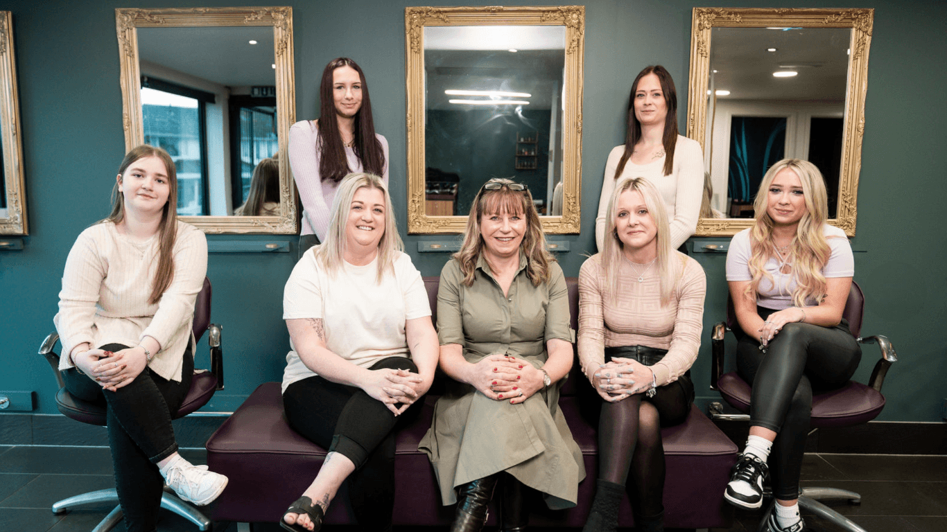 The Honeys Hair & Beauty team pictured together inside the salon in Taunton. Seven smiling team members pose in front of stylish gold-framed mirrors, dressed in soft, neutral tones. Their warm, professional presence reflects the friendly and expert care clients can expect during every visit