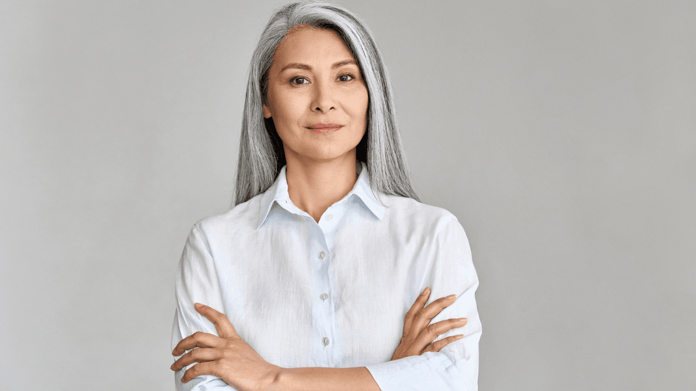 A confident woman with long, beautifully blended silver hair stands against a soft grey background. Her sleek, natural look reflects expert grey blending techniques—showcasing the elegant, seamless results available at Honeys Hair & Beauty in Taunton.
