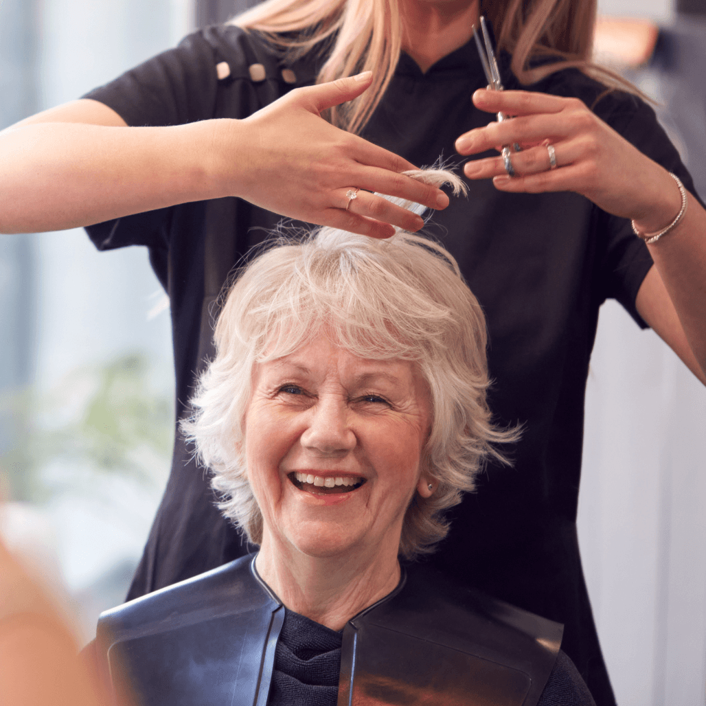 A smiling, silver-haired woman enjoys a haircut at Honeys Hair & Beauty in Taunton. She sits in a salon chair wearing a cape, while a professional stylist trims her hair with scissors. The warm, relaxed atmosphere reflects the expert care and friendly service Honeys is known for, especially for mature clients seeking quality, confidence-boosting styling.