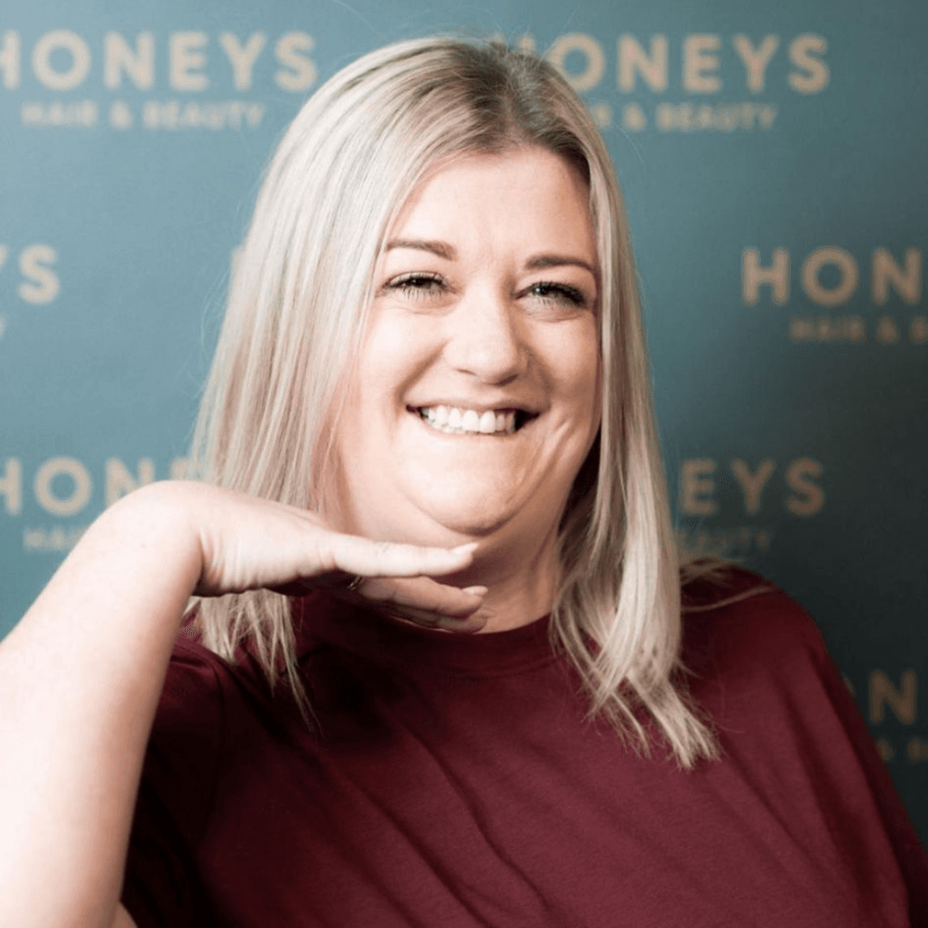 Hannah beams with joy in front of the Honeys Hair & Beauty branded backdrop. Her light blonde hair and playful pose showcase the warm, friendly personality she brings to the salon team in Taunton.
