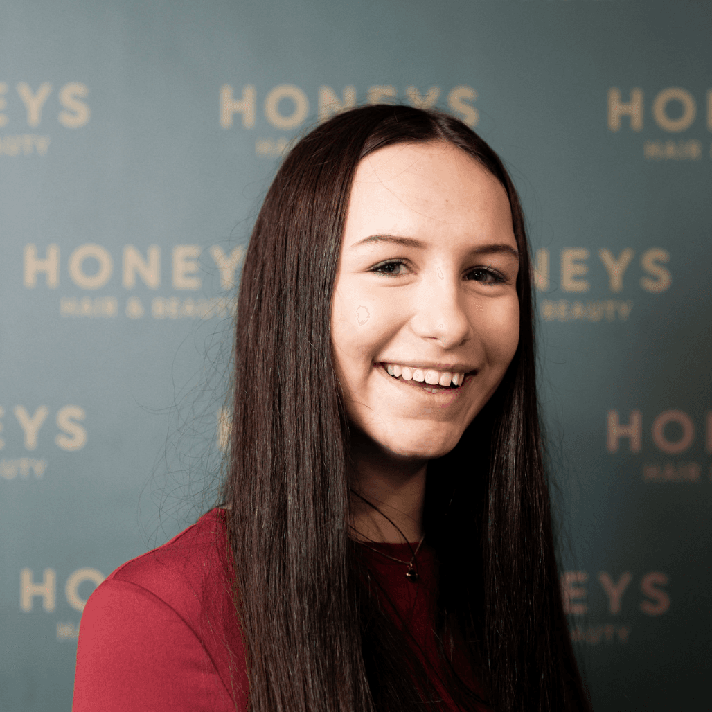 Maddison stands smiling in front of the Honeys Hair & Beauty backdrop. With long, dark hair and a bright personality, she represents the warm, welcoming vibe clients can expect when visiting the salon in Taunton.